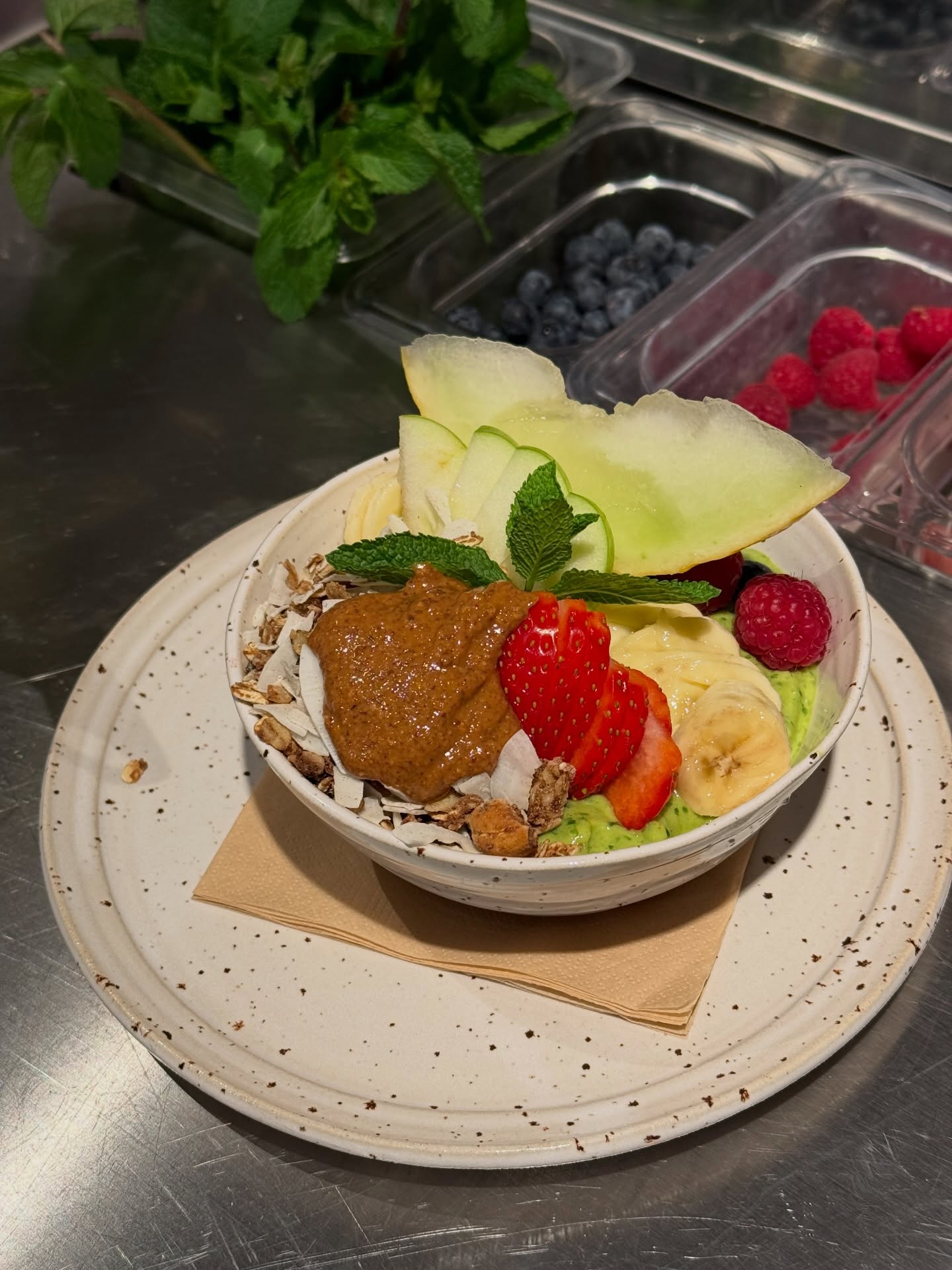A healthy bowl featuring layers of avocado, banana, and strawberries topped with granola, almond butter, raspberries, and green apple slices, garnished with mint leaves.