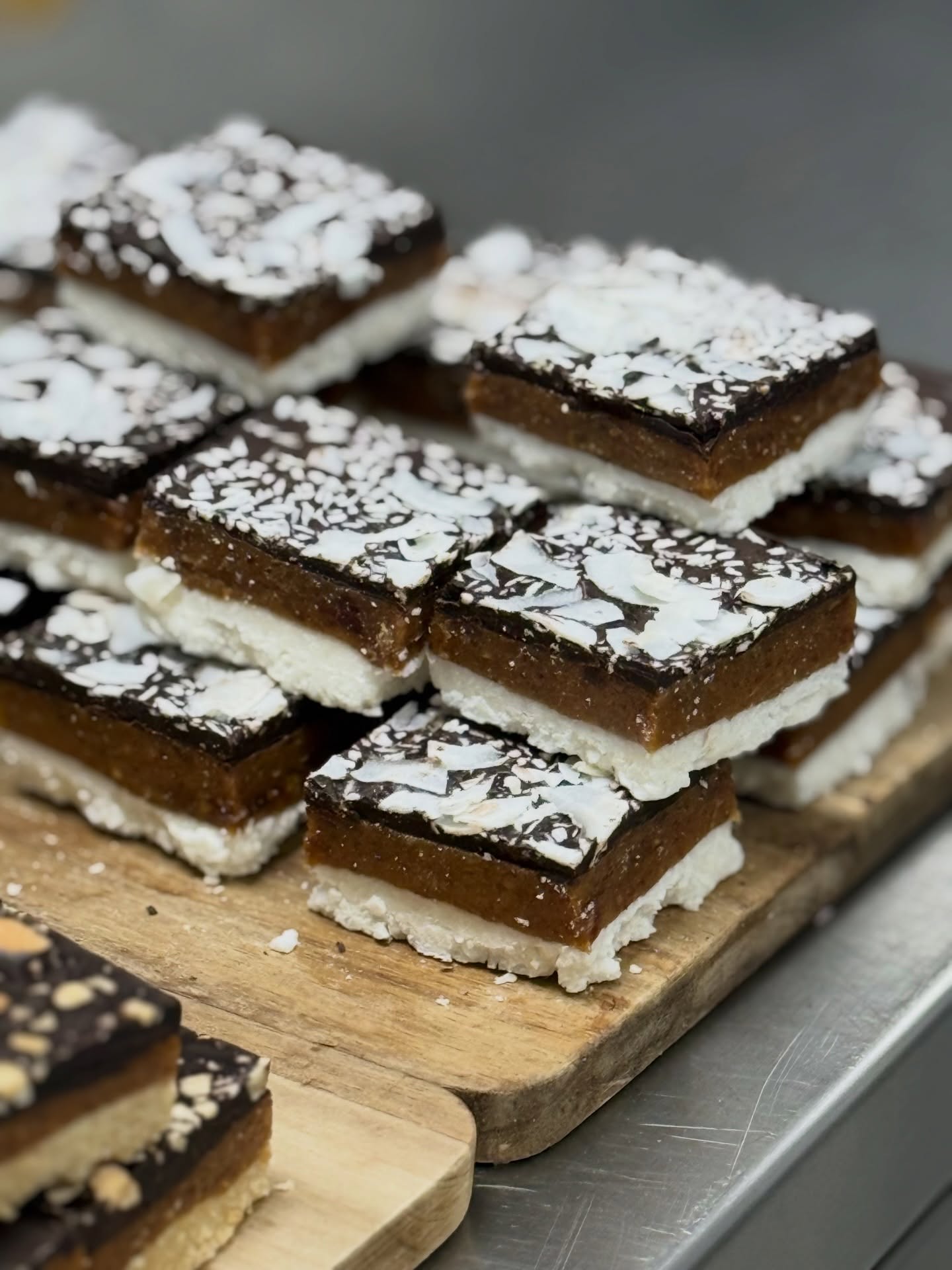 Close-up of stacked square layered dessert bars on wooden boards: a white base, thick caramel-brown middle and glossy dark chocolate top sprinkled with white coconut flakes, blurred kitchen background.