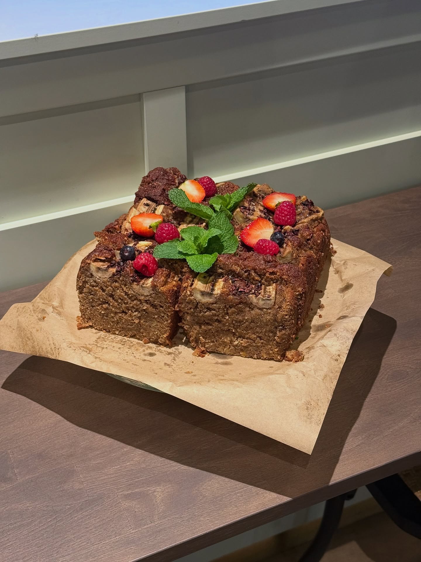 A square cake topped with mint leaves and assorted berries, presented on a piece of parchment paper. The cake has a textured surface and is set on a wooden table with a light-colored wall in the background.