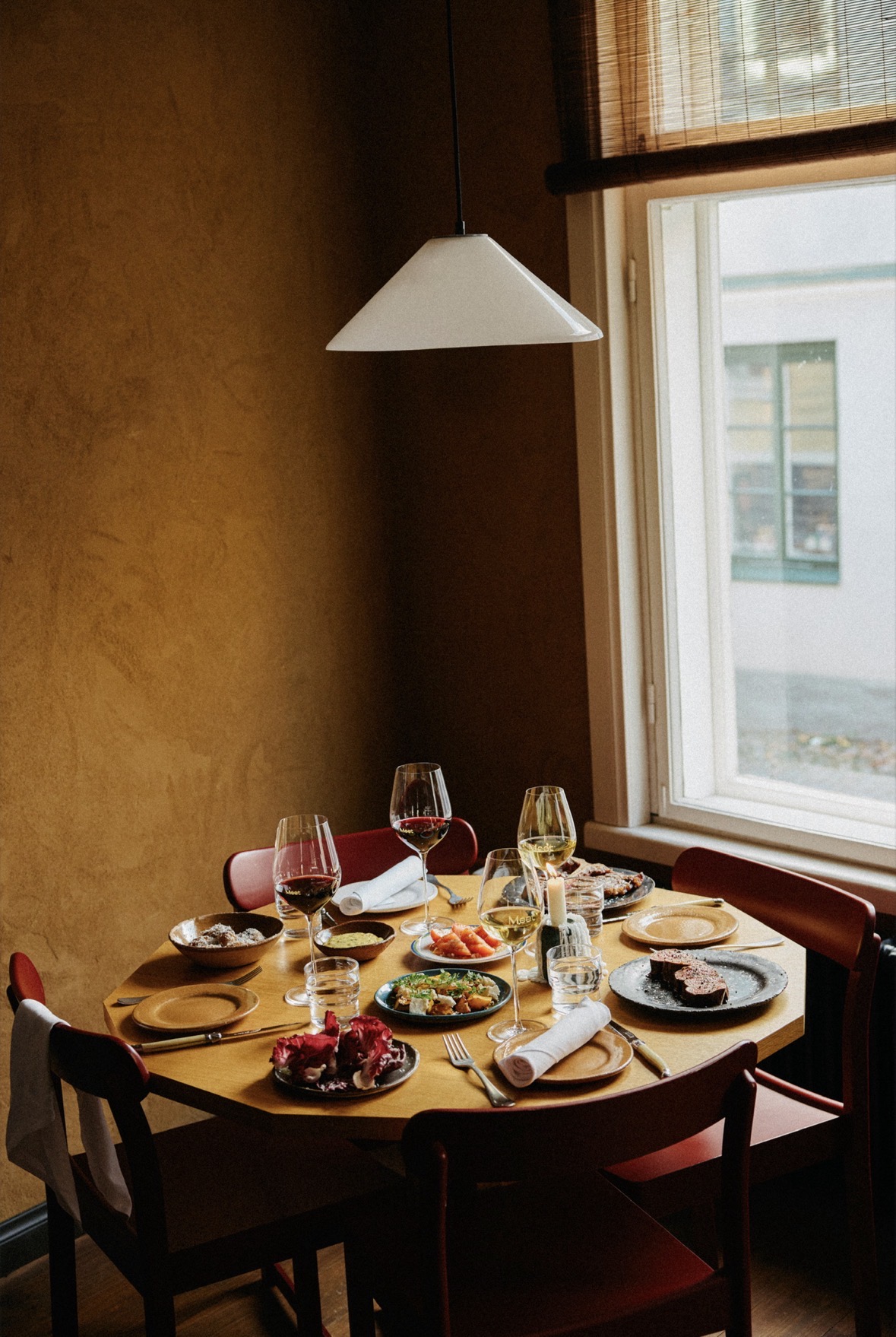 Cozy round dining table set for four beside a window with warm ochre walls and a white pendant lamp. Red chairs, plates, bowls, folded napkins, wine glasses and assorted small dishes.